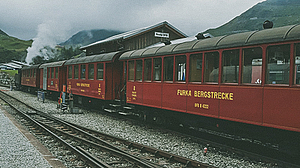 Rote Zugkomposition der Furka-Dampfbahn steht am Bahnhof in Realp vor einem Gebäude, nebelverhangene Berge im Hintergrund.